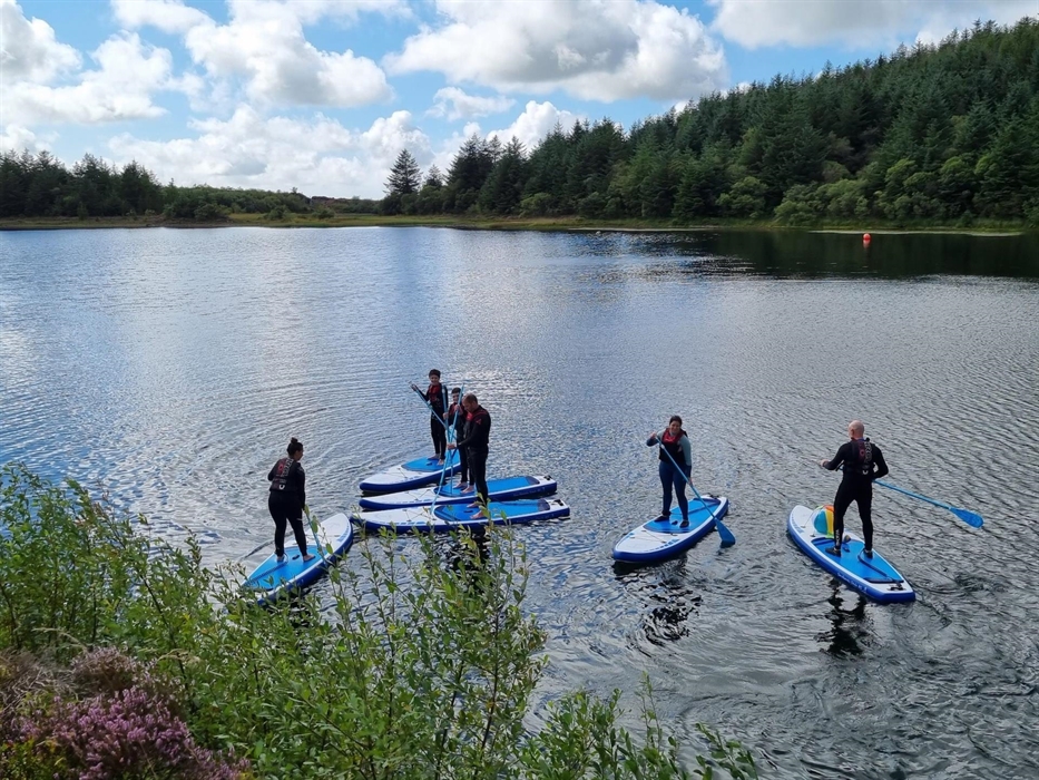A group pof paddle boarders, having a lesson on a lake with trees in tyhe forground and behin the far lake shore.