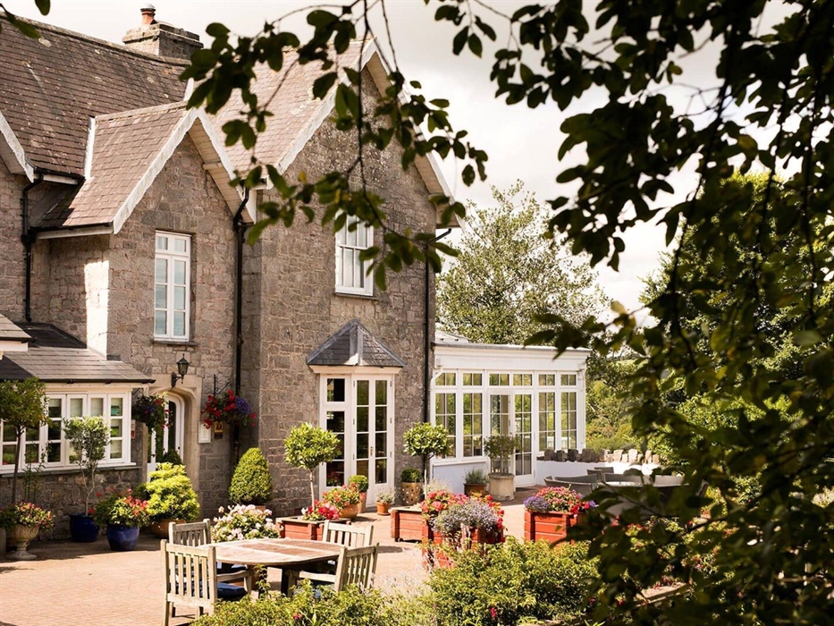 A charming stone house with white-framed windows and a bright conservatory, framed by leafy trees. The patio is filled with colourful flowers, neat to