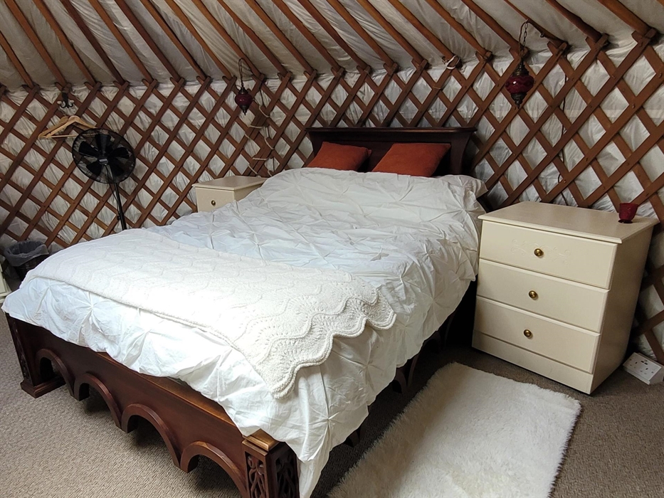 Carved dark wood bed with intricate base details and cream cotton pintuck bedding, cream blanket and burnt orange velvet cushions. Cream painted drawe