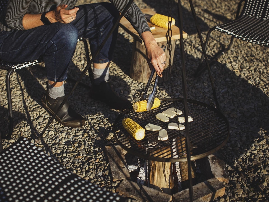 A close up of a campfire scene. A person uses tongs to turn corn on a barbecue hanging over a campfire.