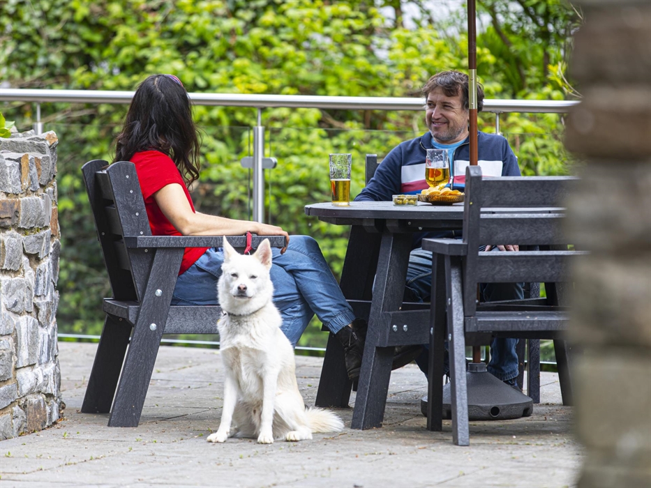 A white dog sits in front of a couple enjoying drinks at a black patio table with stone walls and greenery in the background