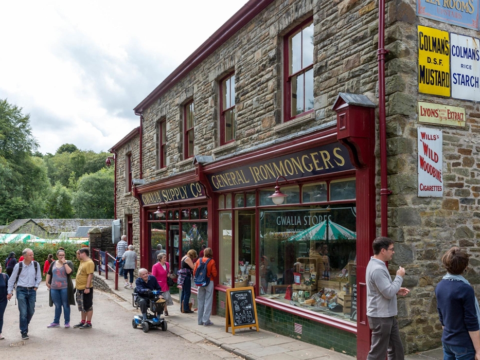 this shows an old shop building made of stone. There are two large shop windows with dark red window frames. you can see Welsh food displayed in one w