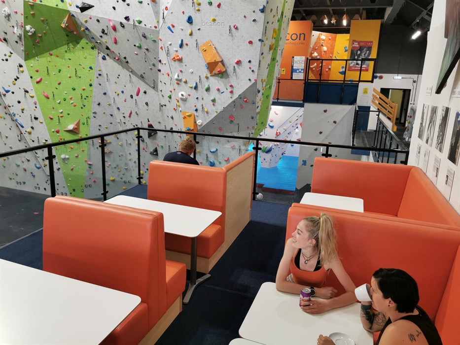Two ladies sit on padded seats at a table in a café area on a balcony overlooking a huge part of the climbing walls.