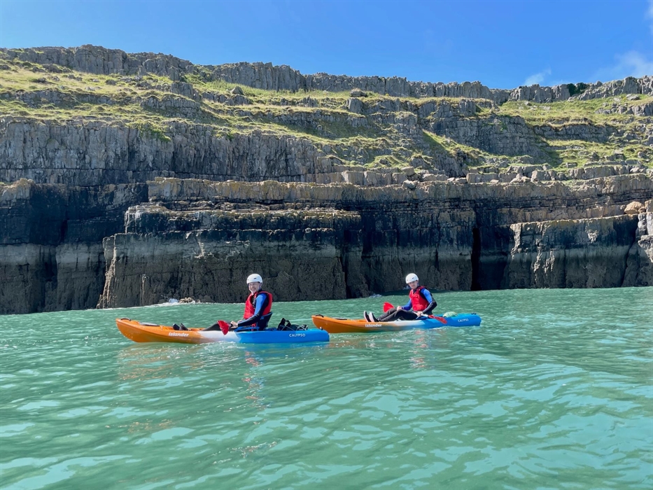 Two kayakers paddling on the sea with dramatic cliffs and clear blue sky in the background