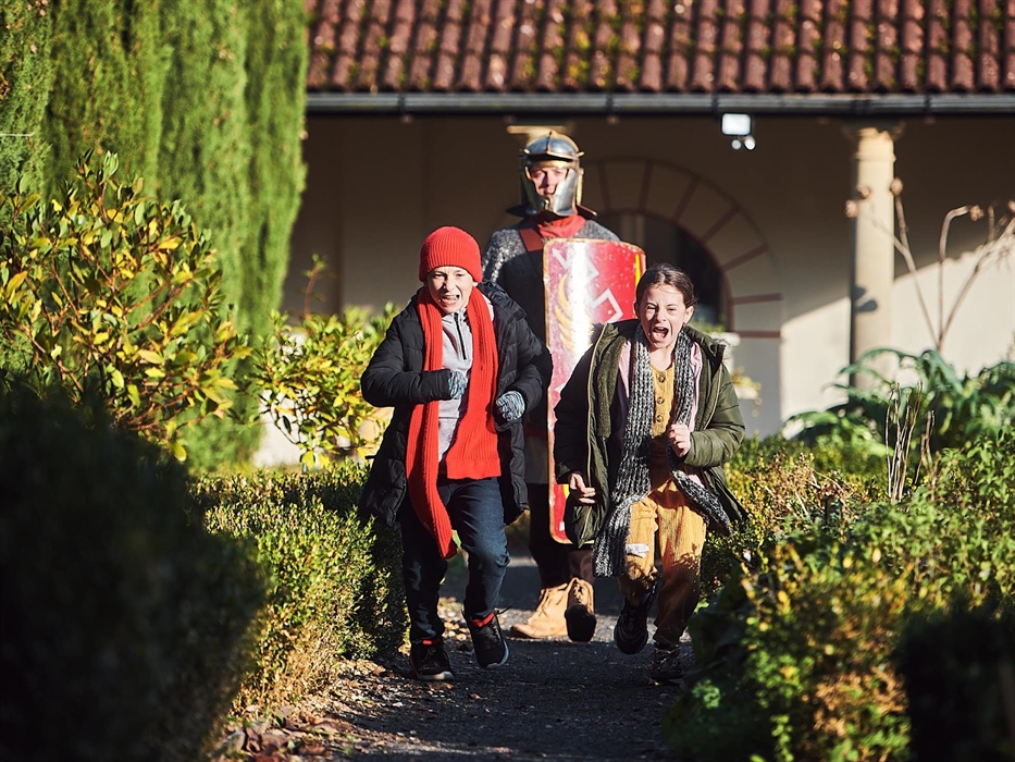 two children run along a gravel path, flanked by low hedges. in the background, there's a man dressed in Roman armour carrying a red shield.