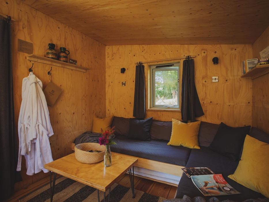 an indoor scene of a cabin with a large corner sofa strewn with scatter cushions and a magazine, a coffee table on a rug sits in the foreground. there