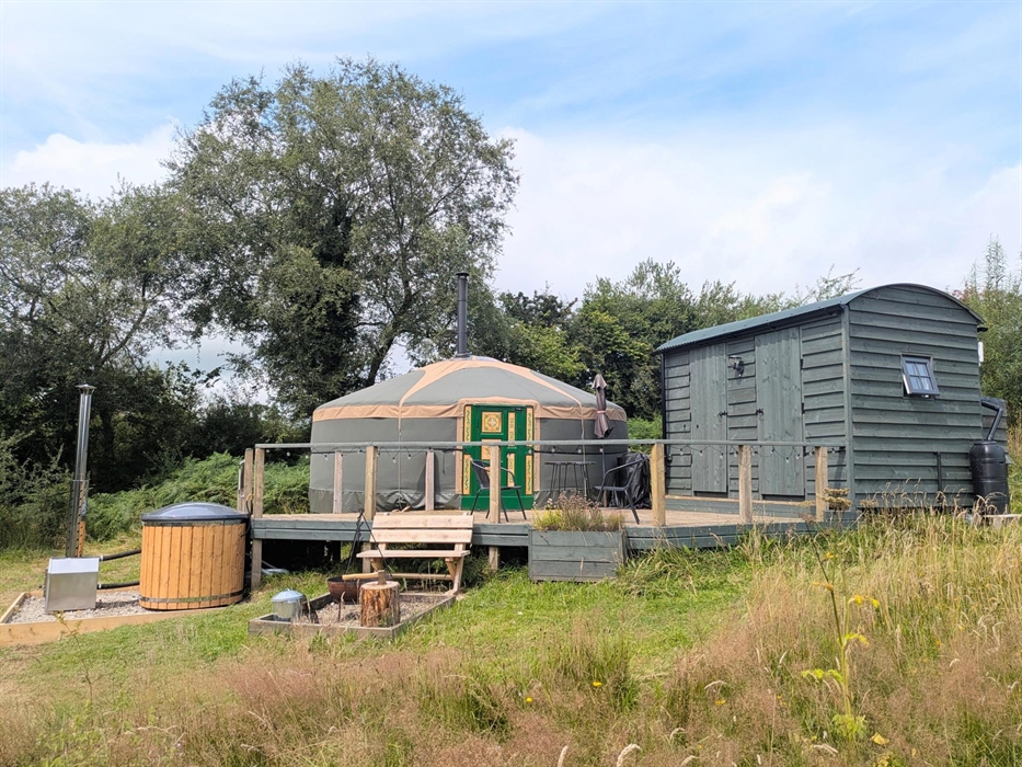A dark green and beige yurt  with a green and pink painted door and a dark green wood hut on a raised deck with a wood hot tub to one side and a fire