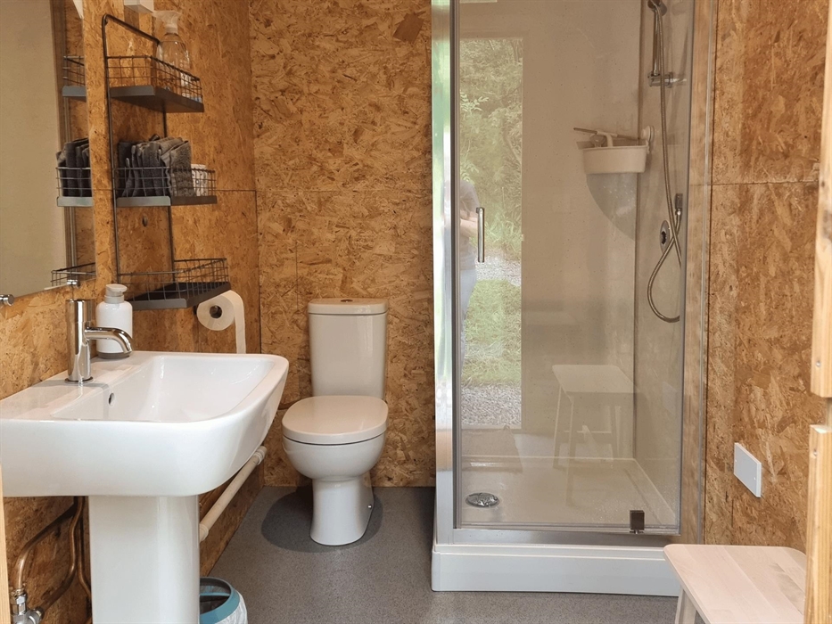 A small room with wooden walls, a shower cubicle, a toilet and wash basin with a mirror above it. There is a wire shelf on the wall