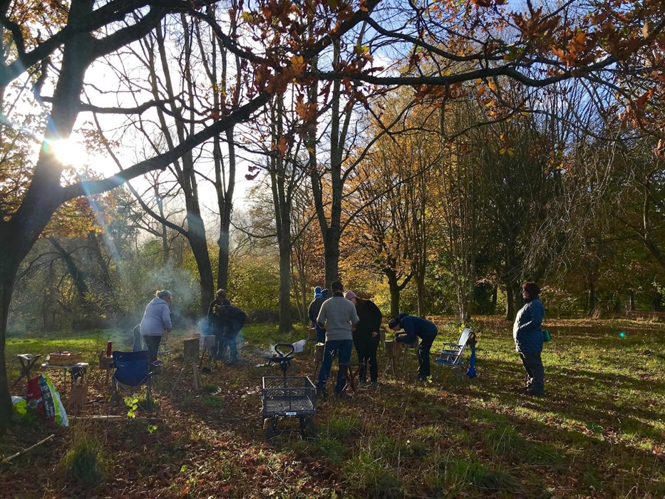 People standing in a circle in a small copse each with their own  2ft upright log as a workstation for carving. Surrounded on left and behind by trees