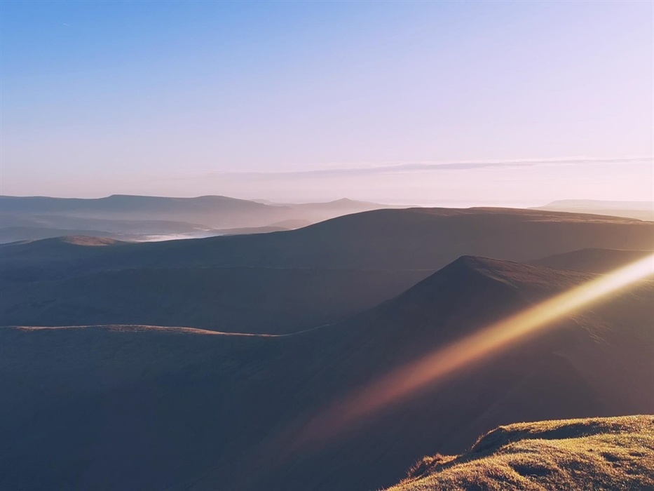 A scene of dawn overlooking layers of mountains of the Brecon Beacons, with golden highlights and pastel tones