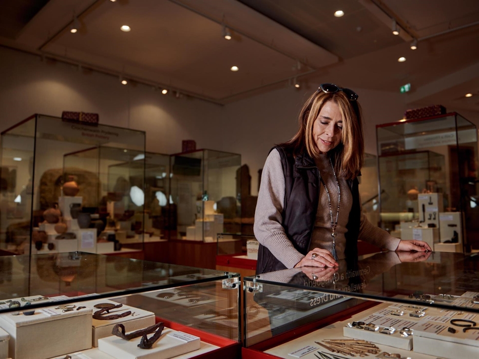 A lady stands over a glass display case. in the case are a selection of metal and other objects found in the archaeological dig at Caerleon