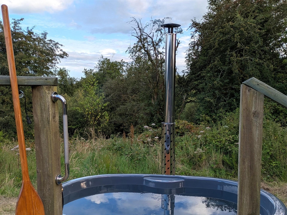 A grey hot tub with chimney, with a wildflower meadow and trees behind