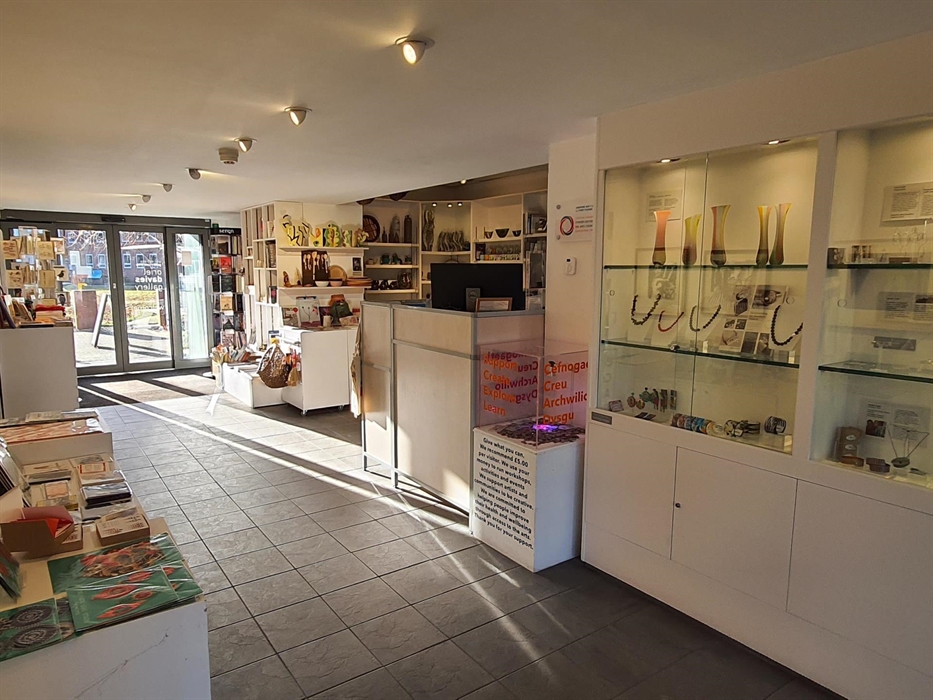 A large glass entrance opens onto tiled floor. Cabinets to the left with cards books and scarfs to the right glass jewelry displays, shelves with bask