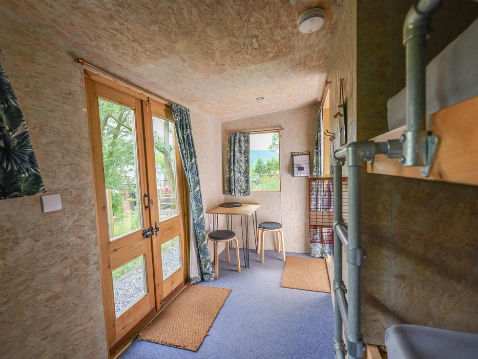 Inside a wooden cabin with double doors and a window, both with curtains. On the floor there is blue carpet, 2 door mats and a small table