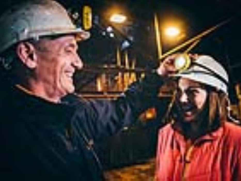 a miner in a hard hat places a white helmet on a lady wearing an orange coat, then switches on the map attached to her helmet