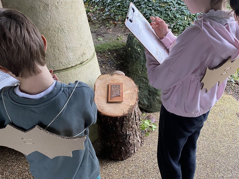 Boy and girl writing on clipboards as part of their outdoor activity. They are also wearing cardboard bats on their back.