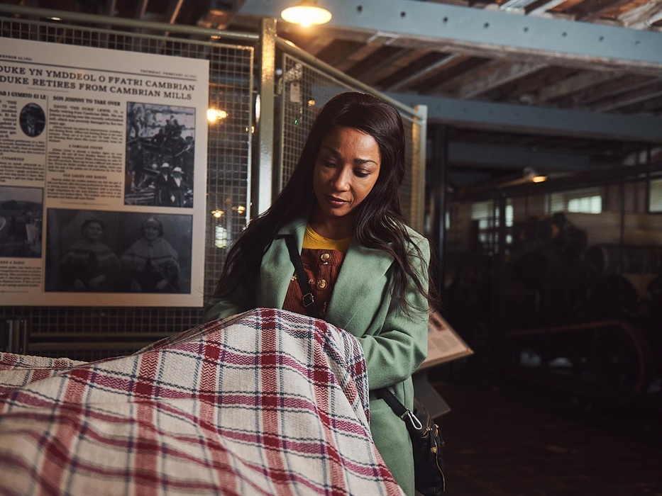 A lady wearing a green coat is lifting up a section of woollen cloth. It is finely woven in a red and cream check pattern, traditional to Cambrian Mil