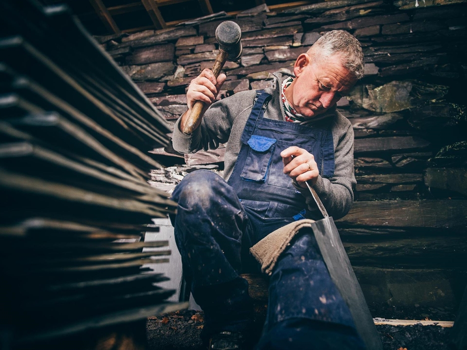 A man in a grey jumper and blue dungarees, sits beside a pile of slates. His right hand is raised, holding a wooden mallet and his left hand holds a c