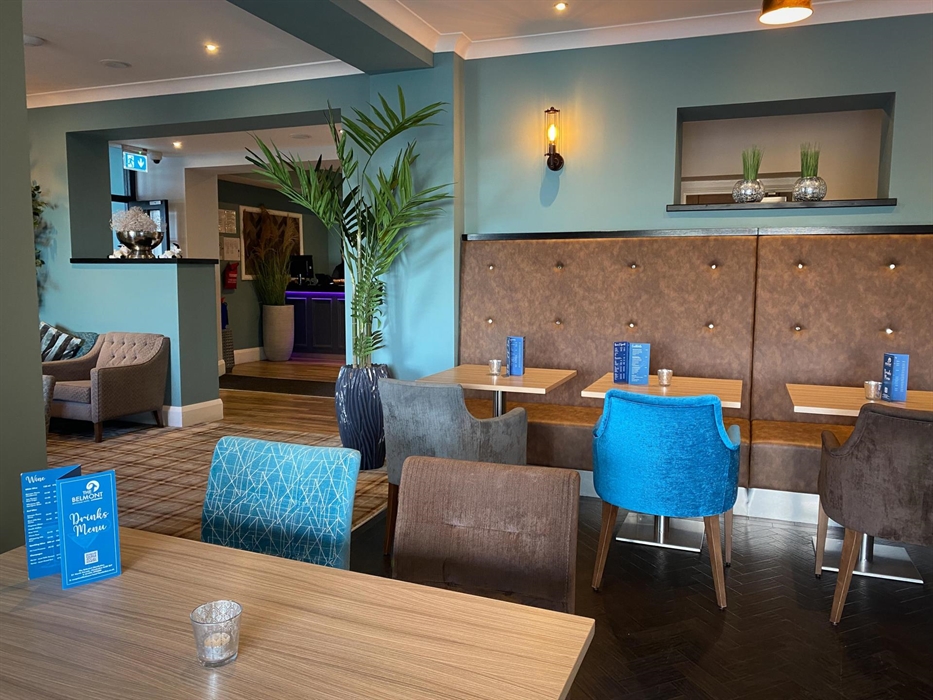 A stylish lounge and dining area featuring a mix of plush seating, wooden tables, and decorative elements. In the background, there is a reception are