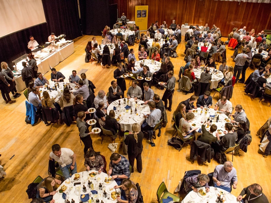 A busy conference dinner, guests being served by staff to round tables.