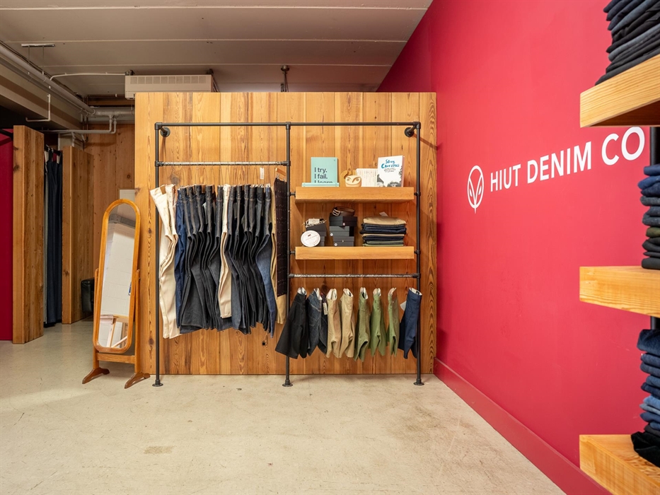Hiut Denim Factory shop display with folded jeans, fabric swatches, and books on wooden shelves against a red wall. A full-length mirror and fitting r