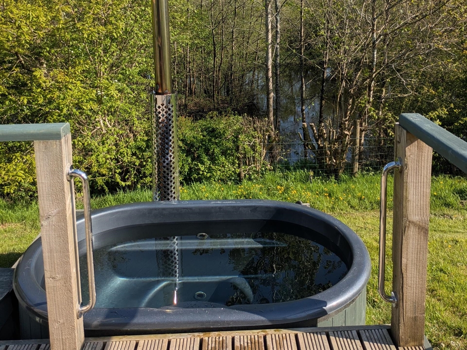 Grey fibreglass hot tub in wood casing next to the wood deck, with a metal chimney behind, overlooking the lake and trees in the background. There are
