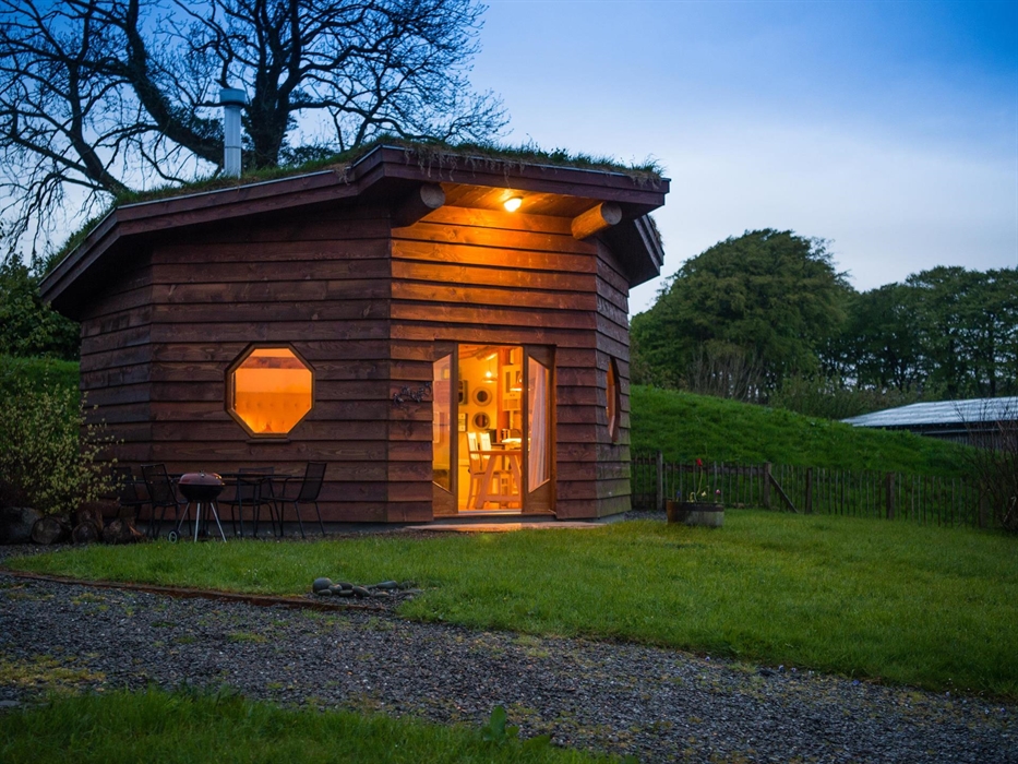 Saffir Lodge sits hidden in a wildflower meadow, its turf roof blending into the hills. Timber walls and warm windows glow beside a steaming wood-fire