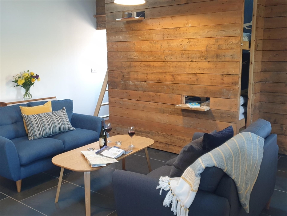 Sofas either side of a coffee table with a map and guide book open and a bottle of wine and 2 glasses.  Bunk beds in the background hidden behind a ti