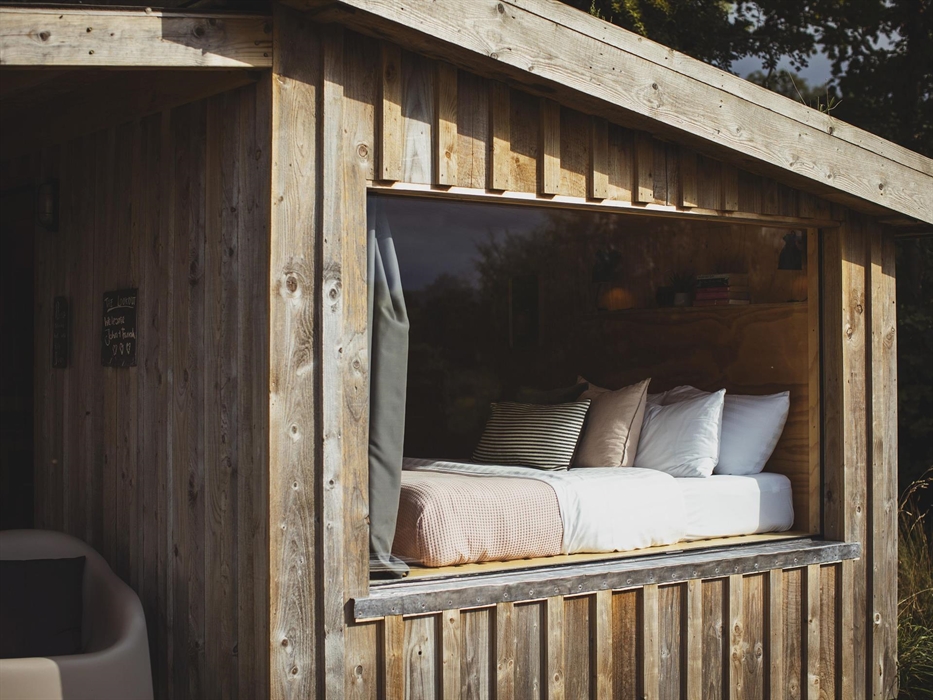 A large picture window looking into a wood cabin with a bed inside with a pink bedspread and scatter cushions.