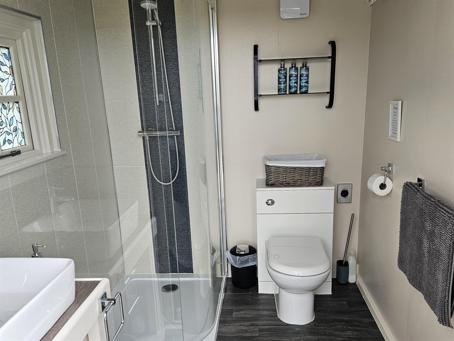 Glass shower cubicle, with cream and charcoal Aquaboard. White toilet and hand basin. Faith in Nature toiletries on black glass shelves. Charcoal viny