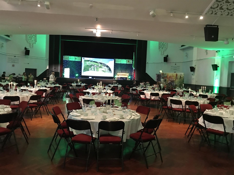 Main Hall Auditorium set up for Awards Dinner, large dressed round tables with place settings and glassware, facing the stage with large screen presen