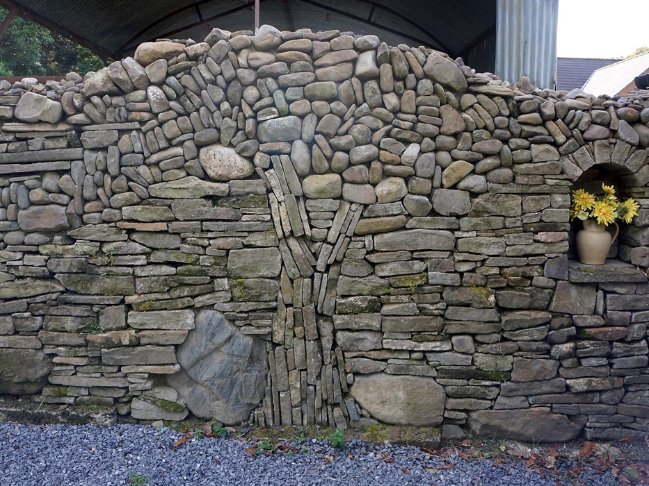 Handcrafted dry stone wall with intricate patterns and a small alcove holding a vase of yellow flowers.