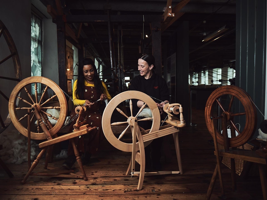 Two ladies sit in front of traditional spinning wheels. One is a craftsperson, showing the visitor how to spin the wool. The visitor slowly feeds the