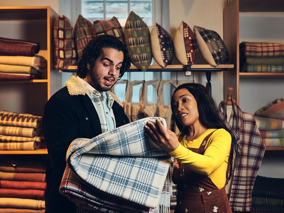 A couple stand in the museum shop. They are holding up a blue and white check blanket, to buy and take home. The blanket was made on the looms at the