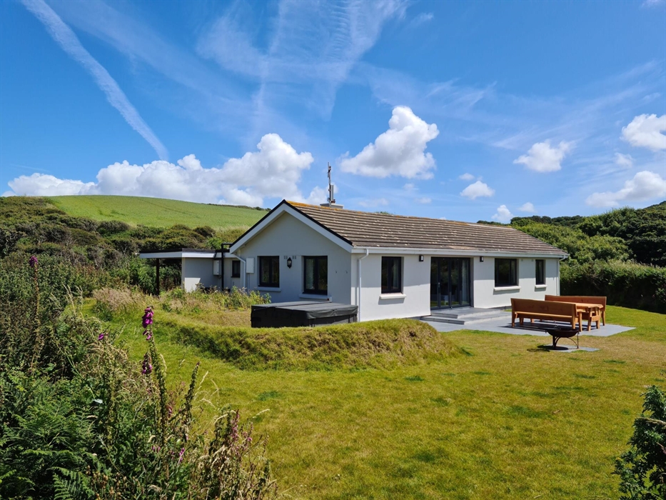 View of Ty'r Felin self catering holiday bungalow and the garden on a sunny day with blue skies. Hot tub, fire pit and wooden table with long wooden b