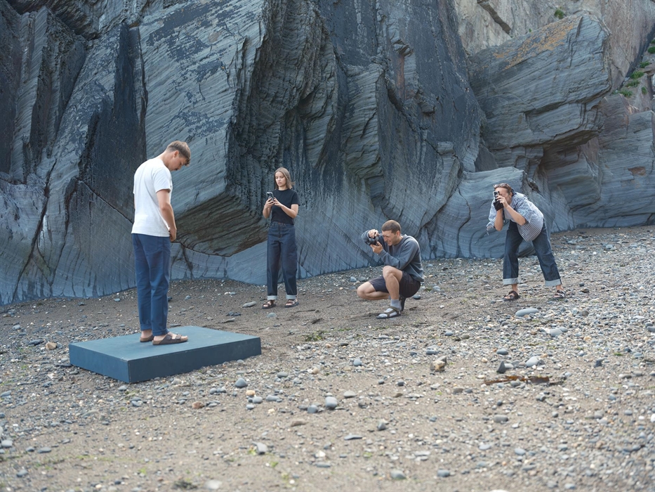 A model stands on a platform by a rocky cliff as three photographers capture images on a pebbled beach. A creative team in casual clothing surrounds t