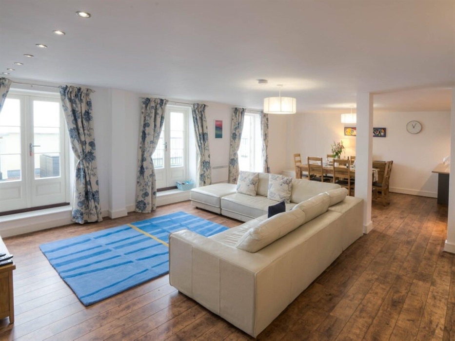 Spacious living room area showing rug, 2 sofas, dining room chairs and table with interior pillar and 3 sets of French doors onto balcony.