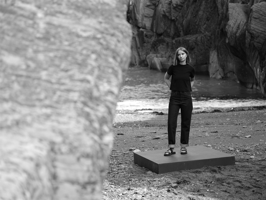 A woman stands confidently on a platform in front of coastal cliffs, wearing a black top and dark jeans. The black-and-white photo emphasises simplici