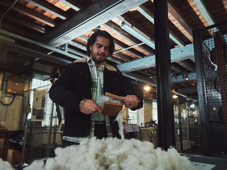 a man stands in front of a pile of washed fleece. he holds two paddles, which resemble large hairbrushes with wire teeth. By scraping a piece of fleec