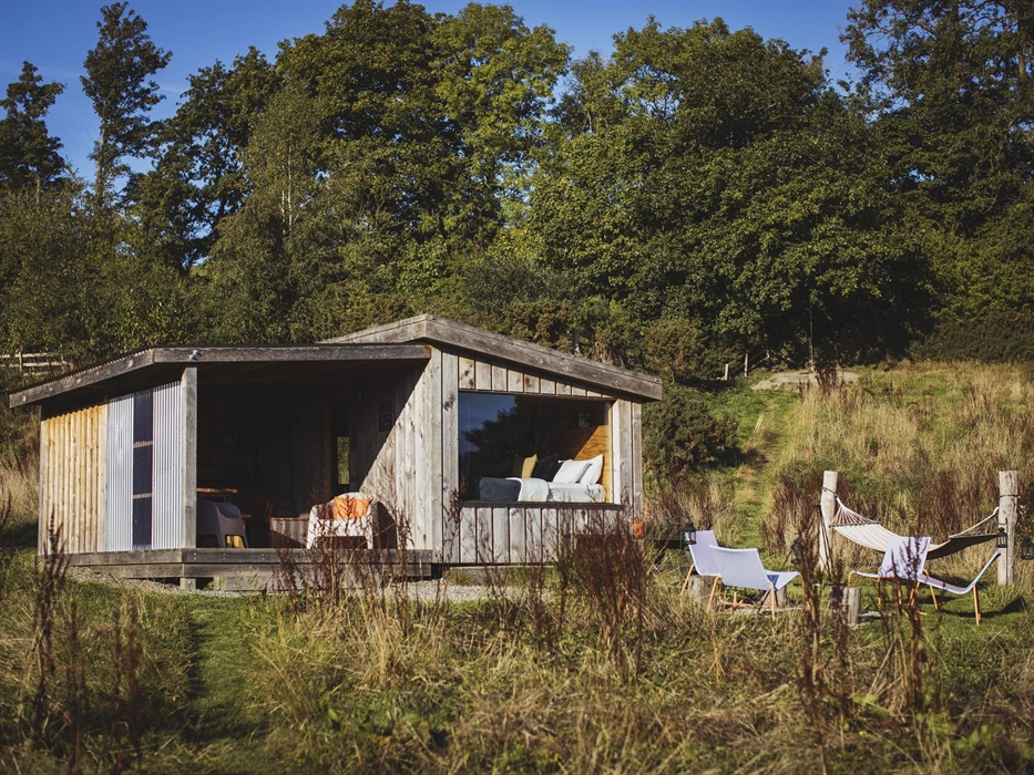 Wood cabin set in a meadow surrounded by trees.  There is large window at the front of the cabin with a bed made up inside. Outdoors is a campfire set