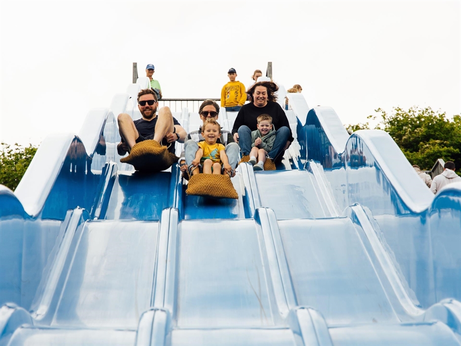3 adults and 2 children sliding down a big blue slide in sacks.