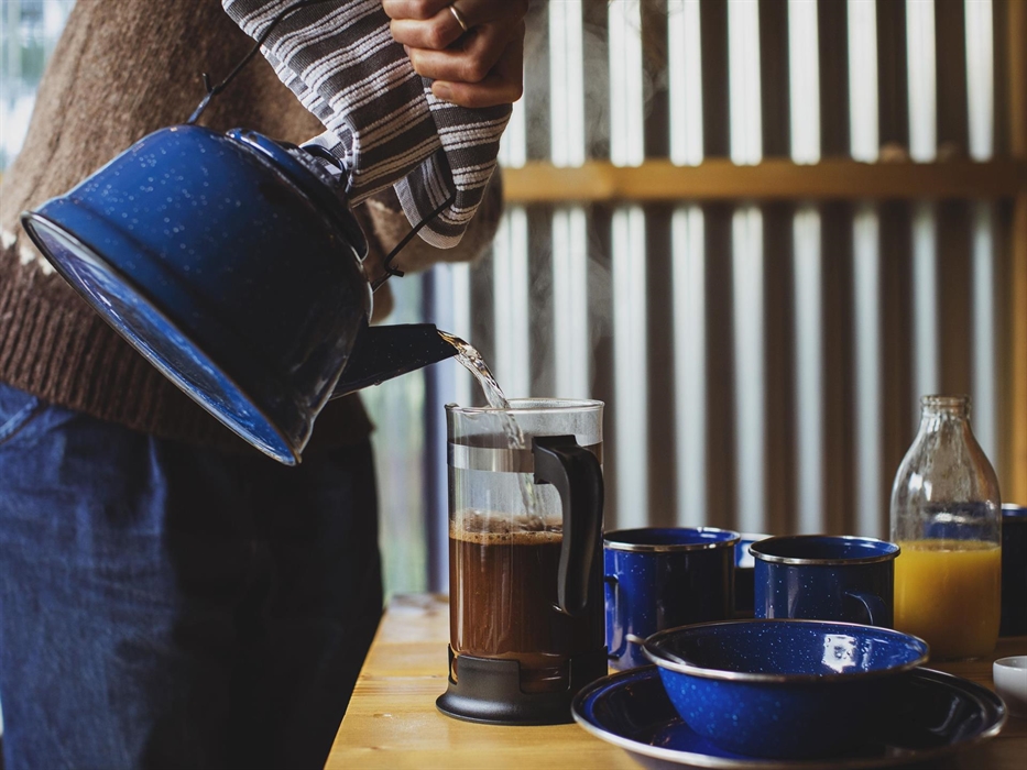 A close up of a person pouring hot water into a cafetière full of coffee. Also a bottle of organise juice and various cutlery.