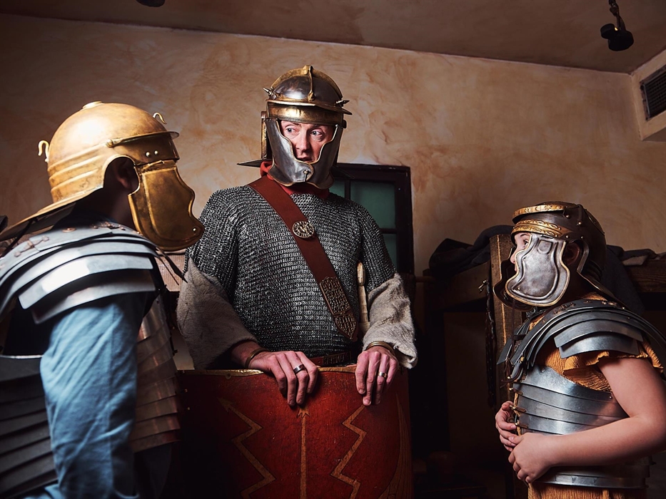 A gladiator stands in the middle of the room. He is flanked by two children trying on Roman armour and helmets. The gladiator explains what it was lik