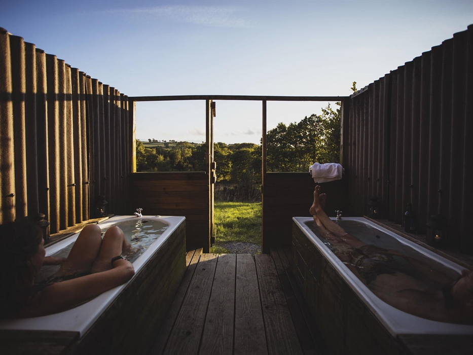 Two outdoor baths sit side by side with a view to the distant landscape of woodland and hills. The baths are full and there is a person soaking in eac