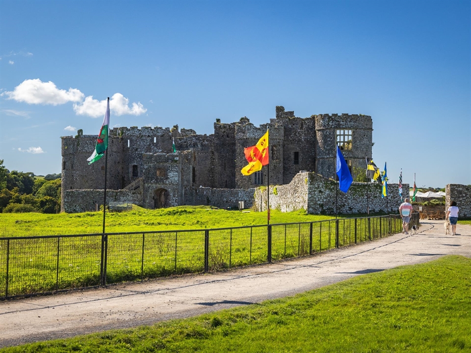 Image of Carew Castle where two dog walkers and walking up the path towards the Castle. Also several types of flags on display on fencing alongside th