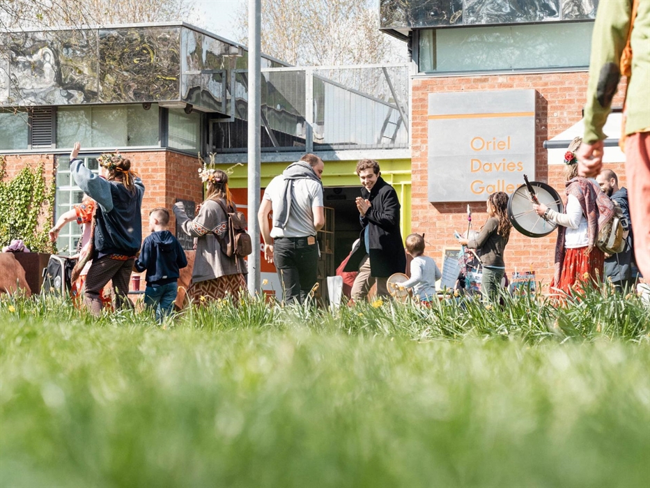 Looking through grass in the foreground Floor level view of people walking in procession in front of the gallery wearing flower crowns carrying and pl