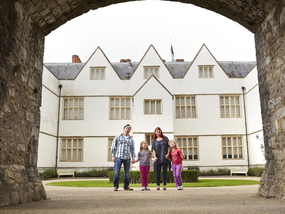 a family of two parents and two young girls stand beneath a high stone archway. in the background is a large white building, with lots of leaded windo