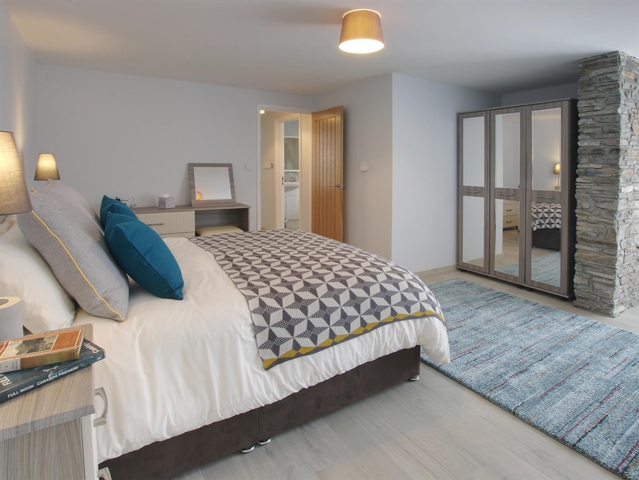 Generously sized master bedroom with modern grey furnishings, a feature stone pillar, and a cosy patterned throw.