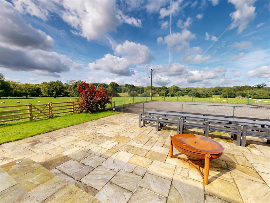 Spacious stone terrace with fire pit, overlooking a fenced tennis court and rolling green fields with grazing sheep in the Welsh countryside.