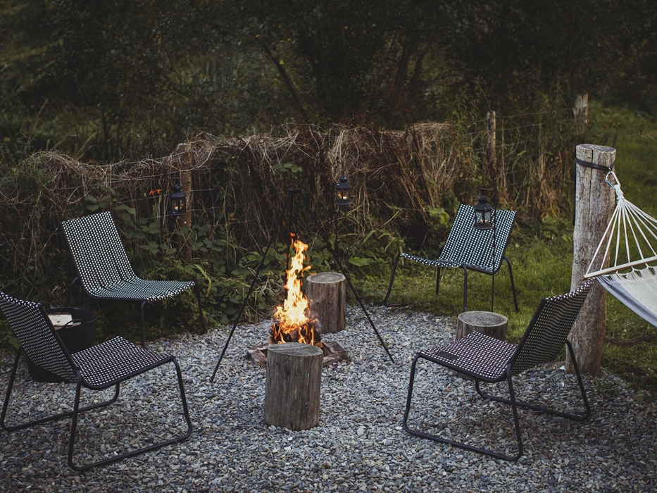 A roaring campfire surrounded by camp chairs set in a rural scene. There is a hammock to one side.